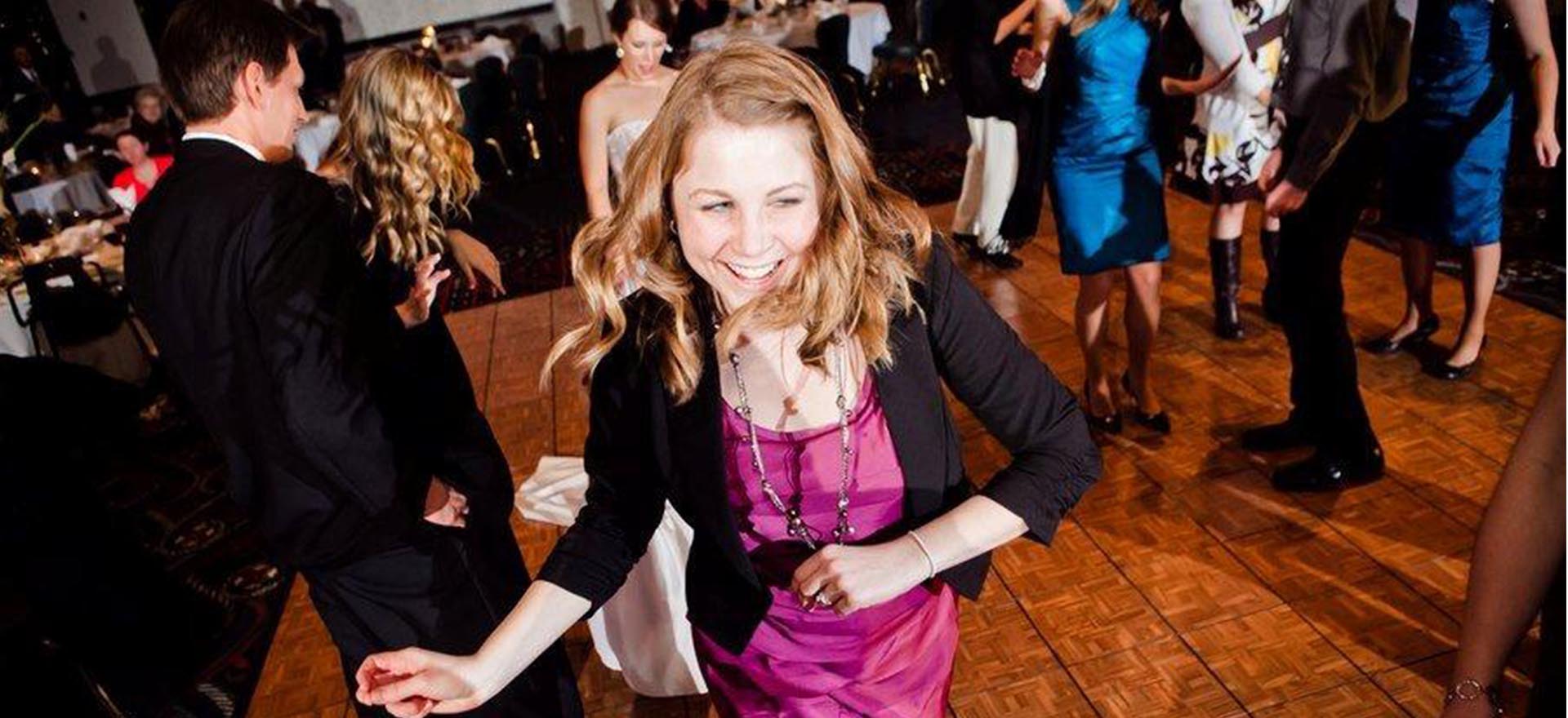 Girl in magenta colored dress and small black jacket with a huge smile in the middle of the dance floor.