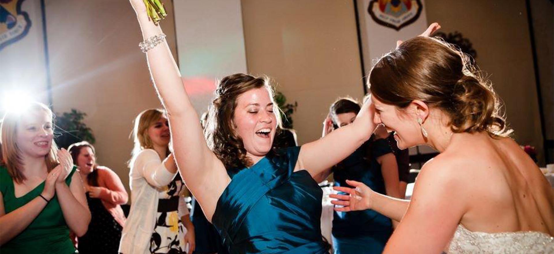 Girl in blue dress with arms in the air dancing and holding a bouquet about to get a hug from her friend in white dress.