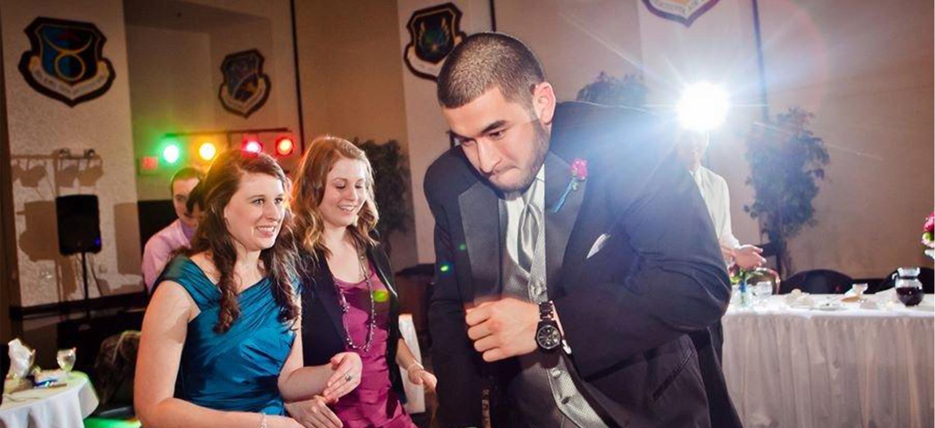 Young man in tux doing a silly 'jogging' dance with girl in blue dress and girl in magenta dressing looking on cautiously.